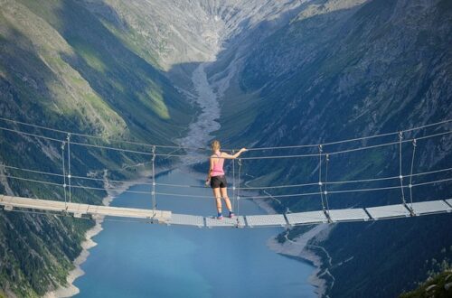 Eine Hängebrücke spannt sich über einen See, der von majestätischen Bergen umgeben ist. Die Brücke ist aus Holz und Seilen gefertigt, und ihre Struktur spiegelt sich im ruhigen Wasser des Sees. Im Hintergrund erheben sich die Berge mit ihren üppig bewaldeten Hängen und steilen Klippen. Der Himmel ist klar und blau, was die idyllische und friedliche Naturkulisse unterstreicht.