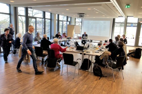 Ein heller, moderner Konferenzraum mit großen Fenstern und Parkettboden. Mehrere Personen sitzen an einem u-förmig angeordneten Tisch und nehmen an einer Besprechung oder Fortbildung teil. Auf einer Leinwand ist eine Präsentation mit einem Schaubild zu sehen. Im Vordergrund geht ein Mann am Tisch entlang, weitere Teilnehmende sitzen oder stehen im Raum. Auf den Tischen stehen Getränke und liegen Laptops sowie Unterlagen.