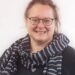 Porträtfoto von Tatjana Leidig, einer lächelnden Person mit Brille, dunklem Oberteil und gestreiftem Schal. Der Hintergrund ist neutral weiß gehalten.