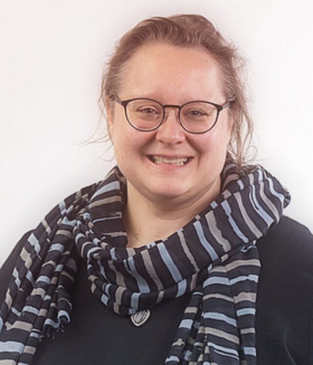 Porträtfoto von Tatjana Leidig, einer lächelnden Person mit Brille, dunklem Oberteil und gestreiftem Schal. Der Hintergrund ist neutral weiß gehalten.