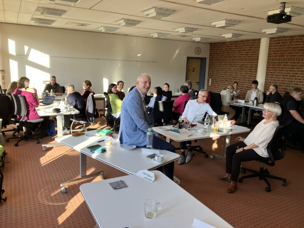 Heller Seminarraum mit mehreren Tischgruppen, an denen Menschen sitzen und diskutieren. Auf den Tischen stehen Laptops, Notizen, Getränke und Tassen. In der Mitte sitzt ein Mann im hellblauen Sakko und lächelt zur Kamera, während die übrigen Teilnehmenden miteinander im Gespräch sind.